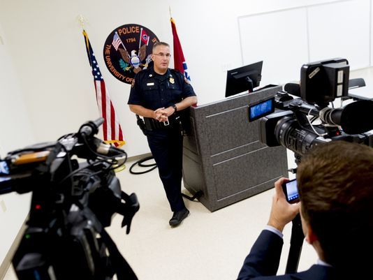 man in police uniform leaning on the desk with tennessee and us flag besides him with cameras pointing to him